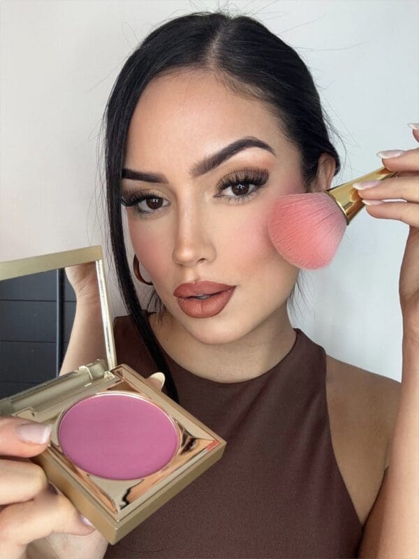 A woman holding a pink blush and wearing brown makeup.